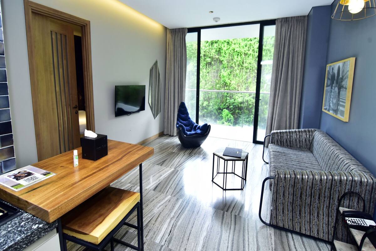 A modern living room with a wooden dining table, a blue bean bag chair, a patterned sofa, and a television on the wall.