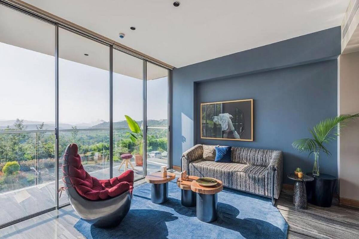 A modern living room with a large glass wall, a red chair, a grey sofa, and a blue rug.