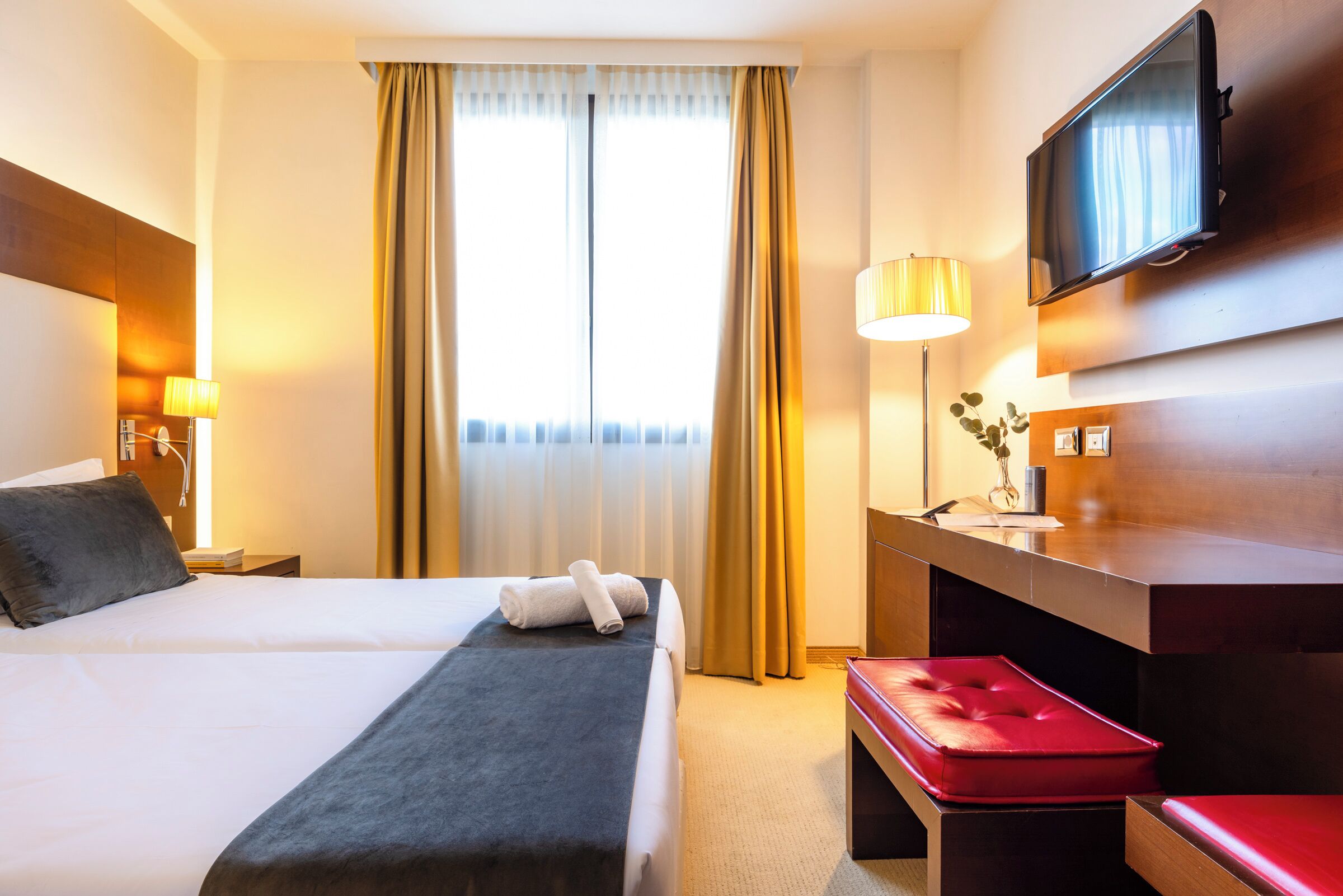 A hotel room with two beds, a TV, a desk, and a red bench.