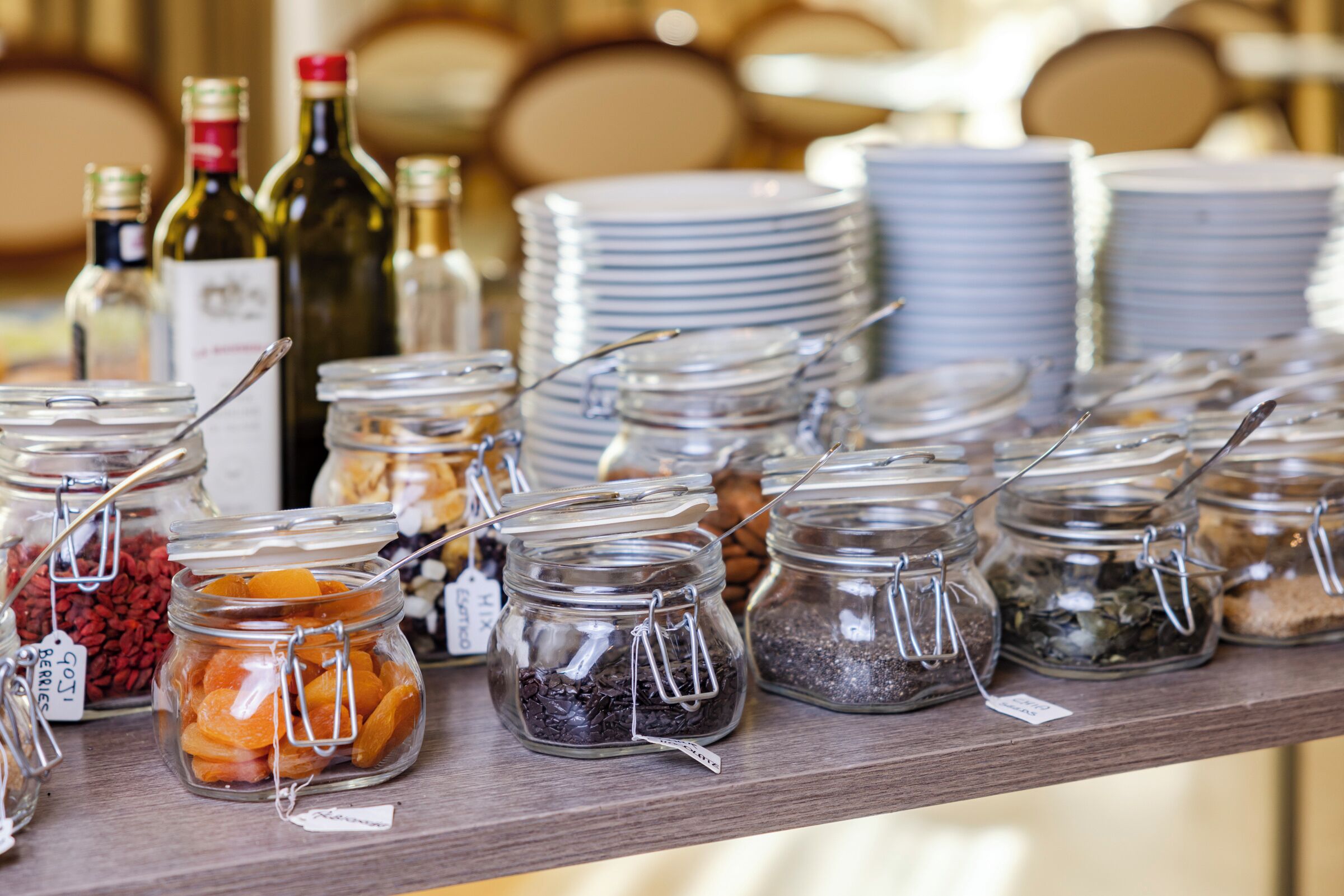 A buffet table with various food items in jars, surrounded by stacks of plates and wine bottles.