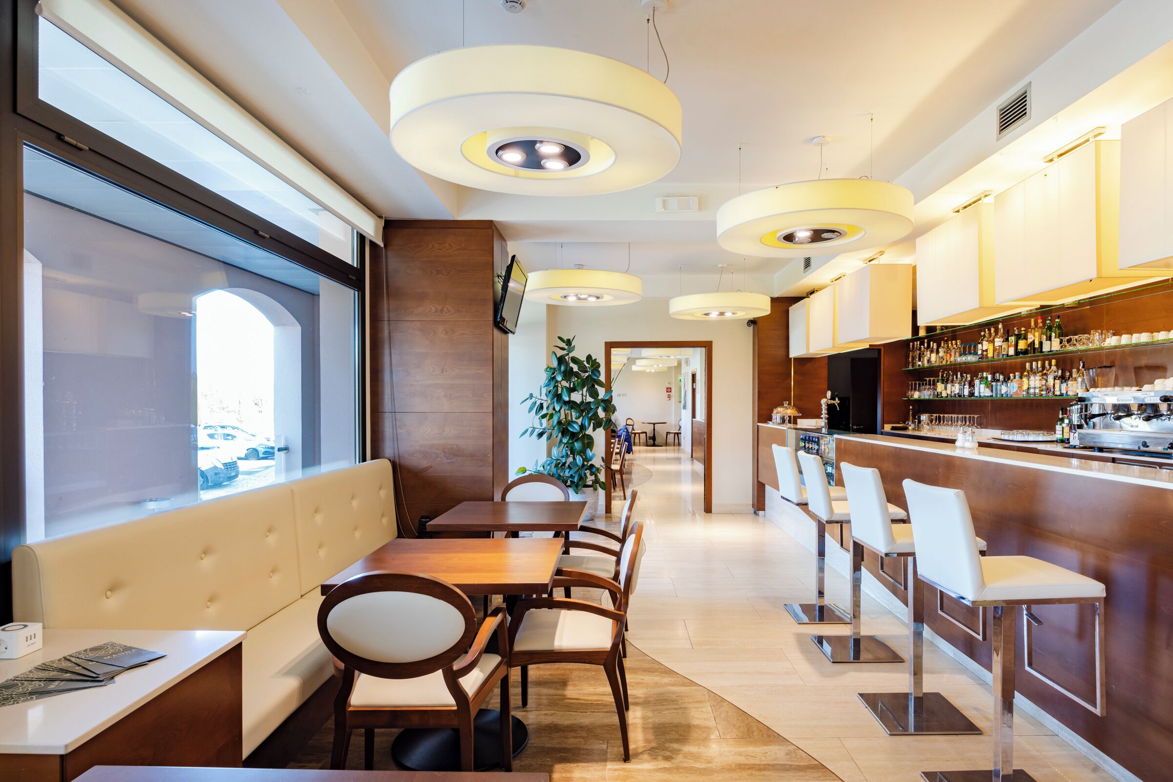 Un ristorante moderno con zona bar, tavoli e sedie in legno e un'ampia vetrata che offre vista sulla strada.
