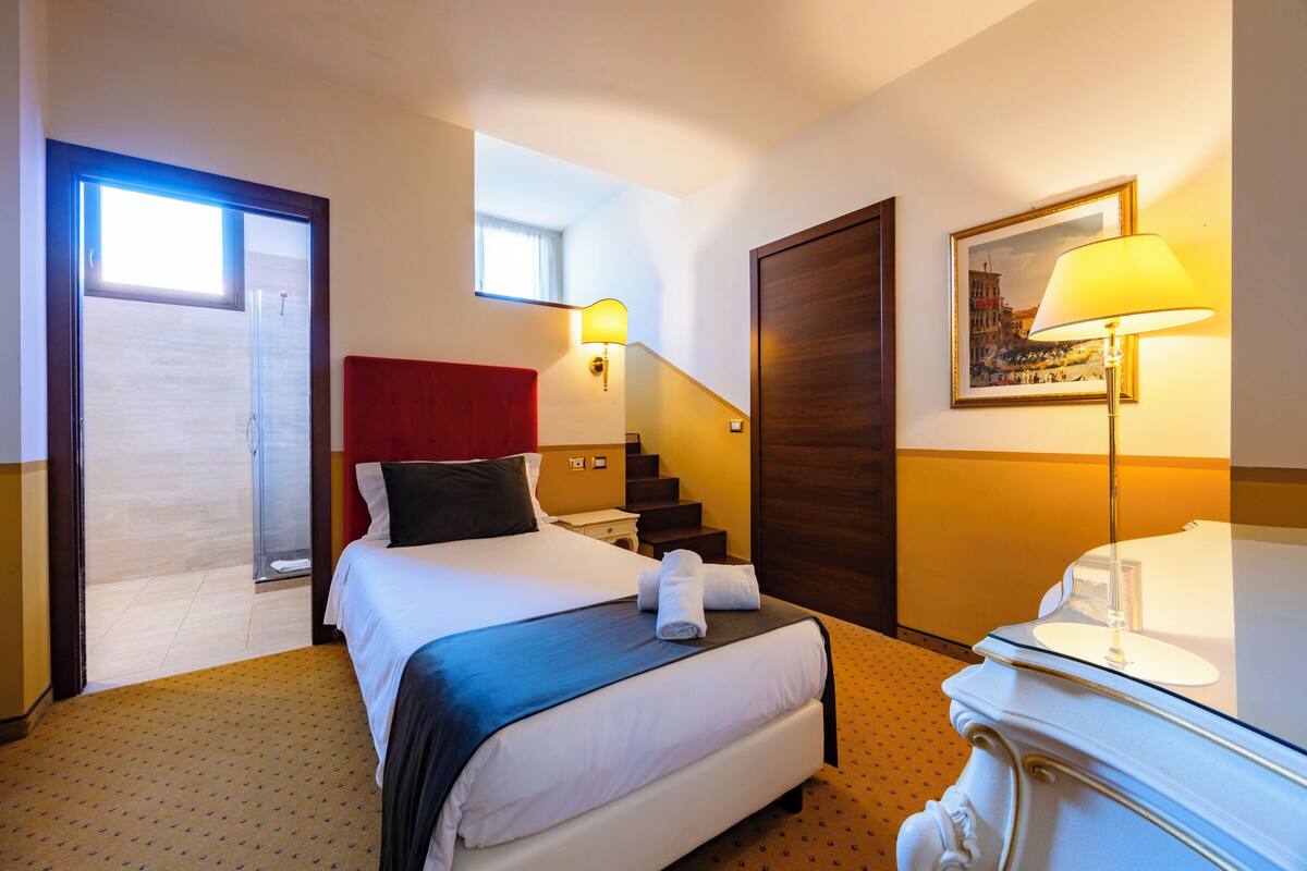 A hotel room with a bed, a nightstand, a lamp, a painting, a door, and a window.