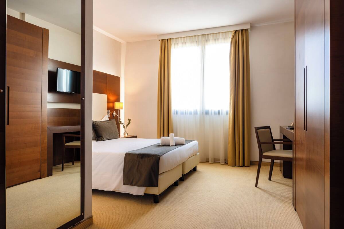 A hotel room with a bed, a desk, a chair, a TV, and a large window with curtains.