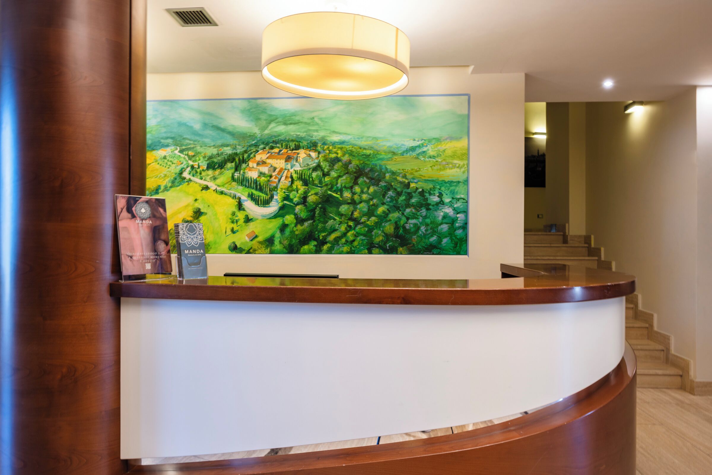 A reception desk with a large painting of a landscape, a modern light fixture, and a staircase in the background.