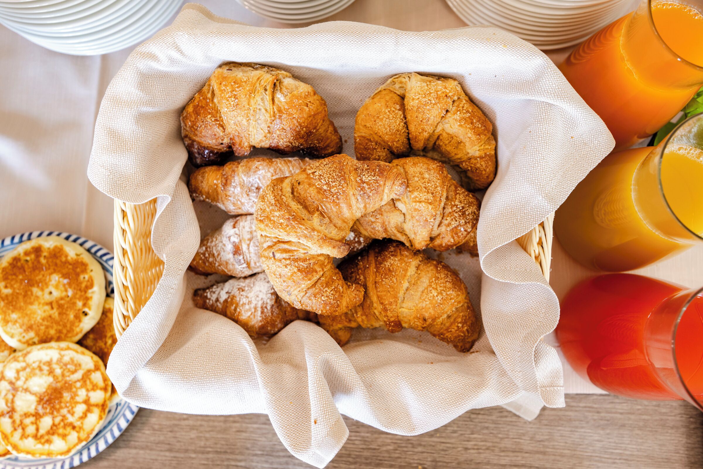 Un cestino con croissant e pancake su un tavolo, insieme a bicchieri di succo d'arancia.