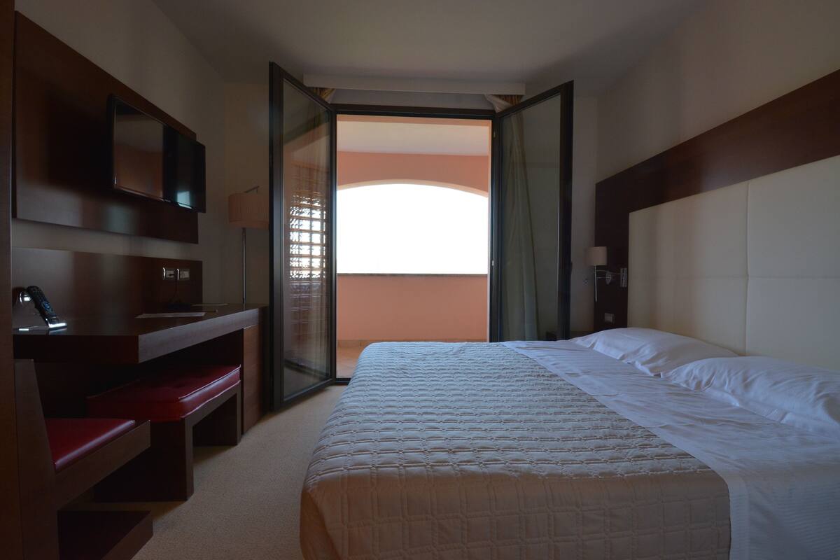 A hotel room with a bed, a desk with a chair, a television, and a large window.