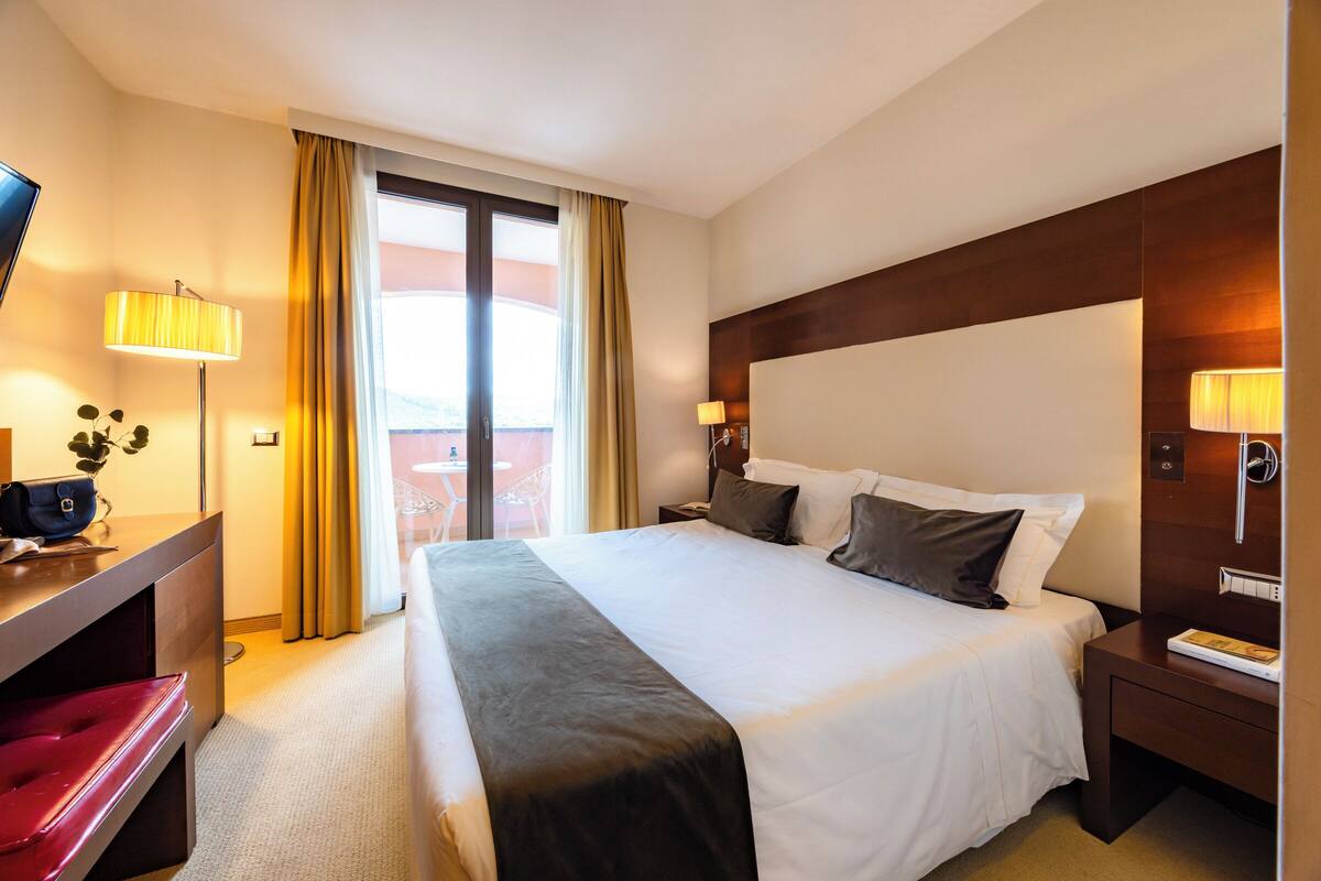 A hotel room with a bed, bedside lamps, a desk with a chair, and a view through a large window.