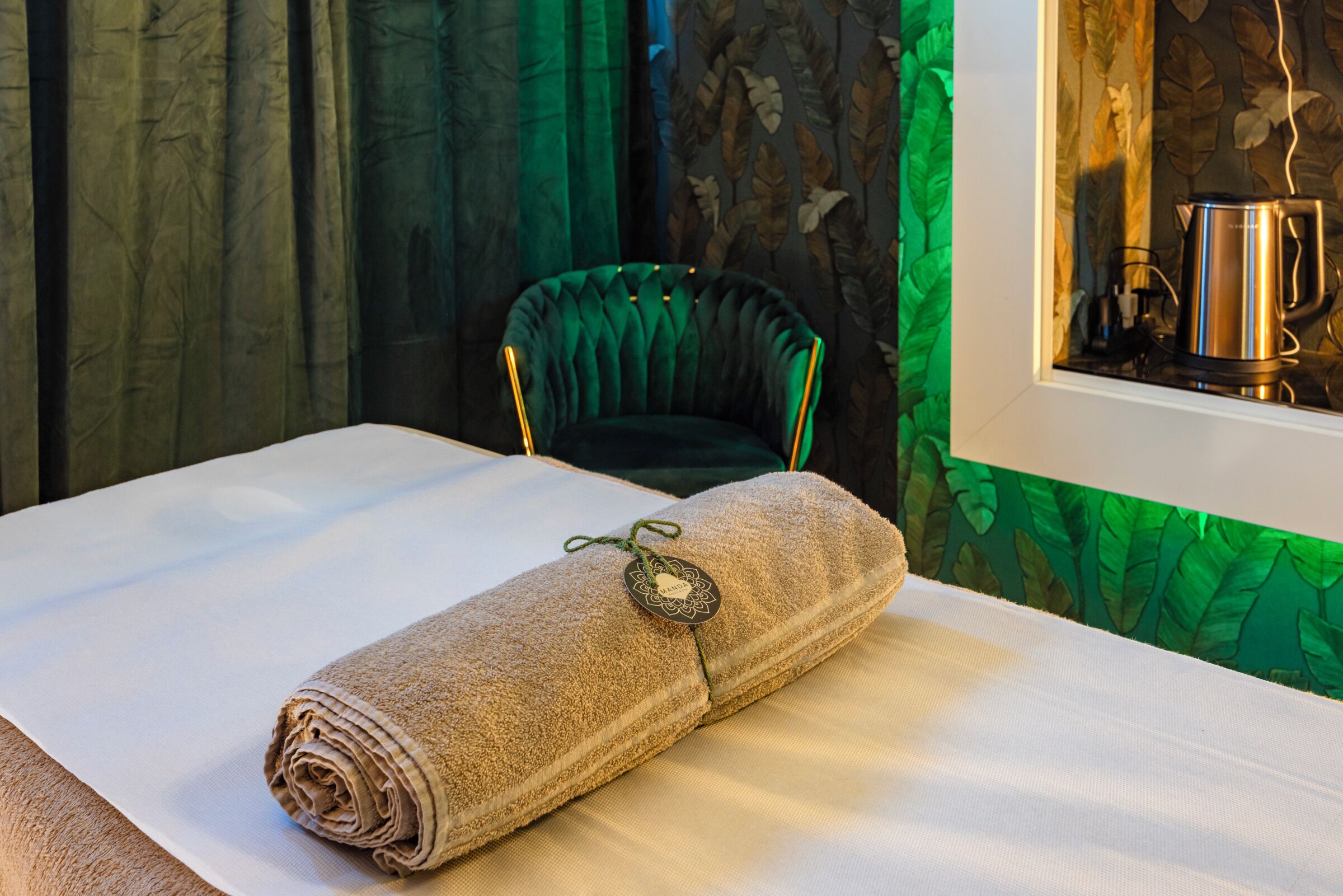 A neatly made bed with a rolled towel and a decorative tag, a green upholstered chair, and a window with a kettle.