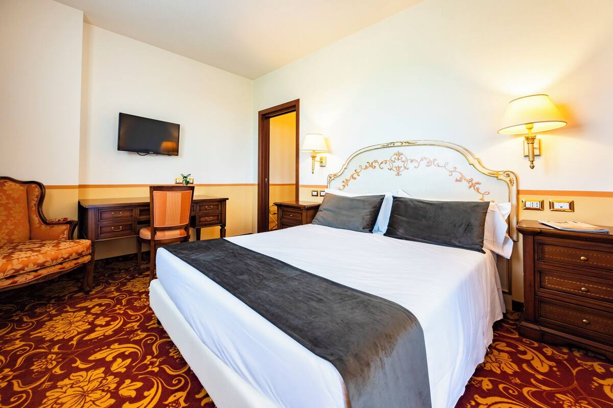 A hotel room with a large bed, a desk with a chair, a television, and two wall-mounted lamps.
