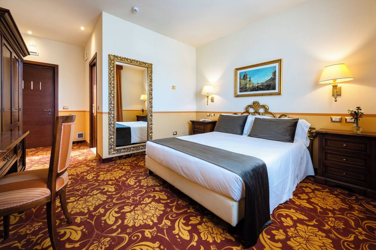A hotel room with a large bed, a mirror, a painting, a desk, and a chair.