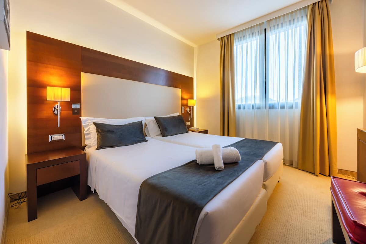 A hotel room with a large bed, two bedside tables, a nightstand, a lamp, and a window with curtains.