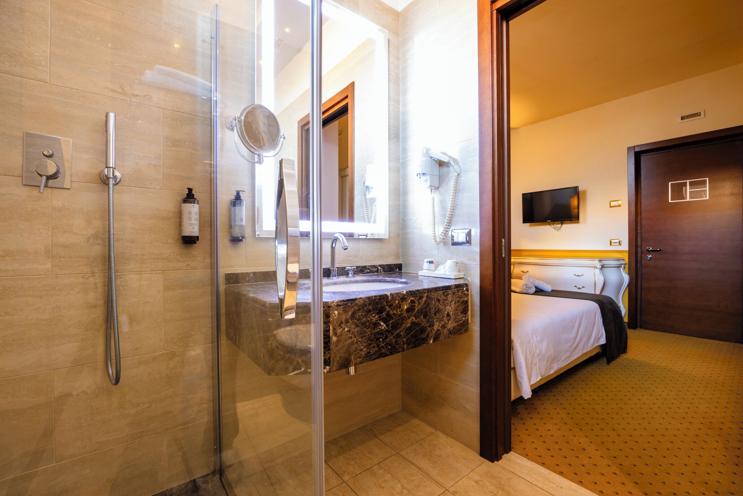 Une salle de bain d’hôtel avec une douche en verre, un lavabo en marbre, un miroir, un sèche-cheveux et un lit avec un téléviseur dans la pièce adjacente.