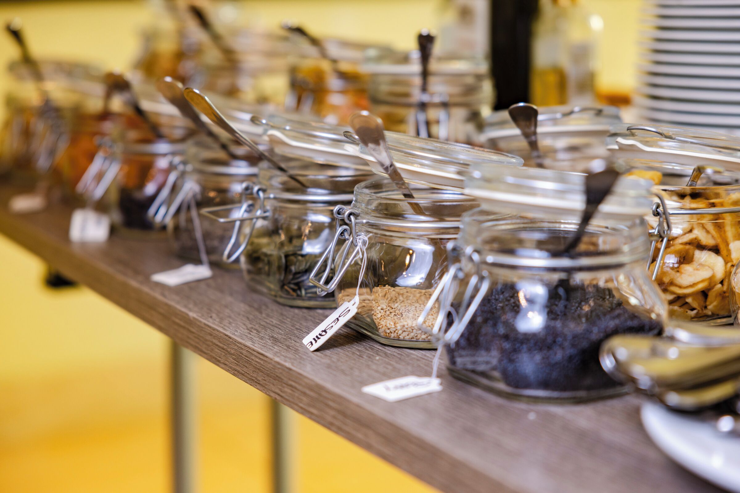 Una barra de buffet con diversos alimentos en recipientes de vidrio, cada uno con una cuchara y etiquetas.