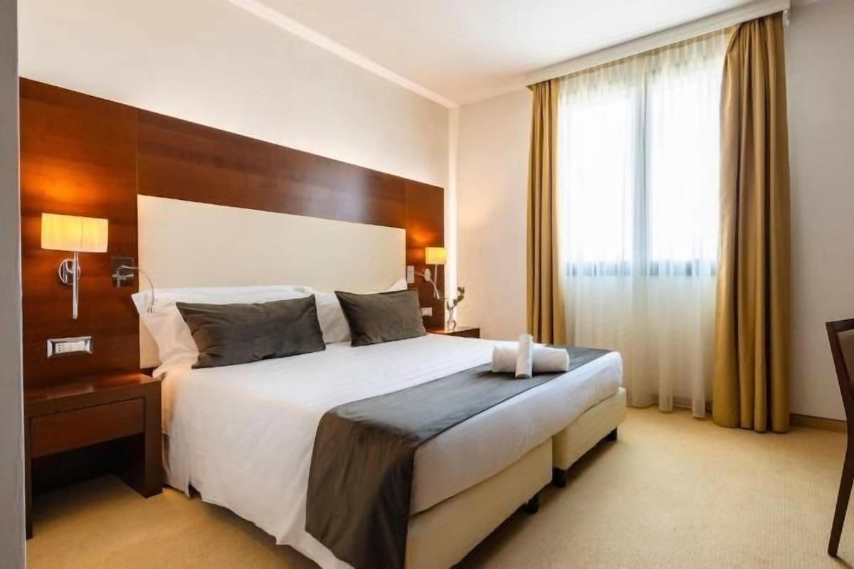 A hotel room with a double bed, bedside tables, a desk with a chair, and a large window with curtains.