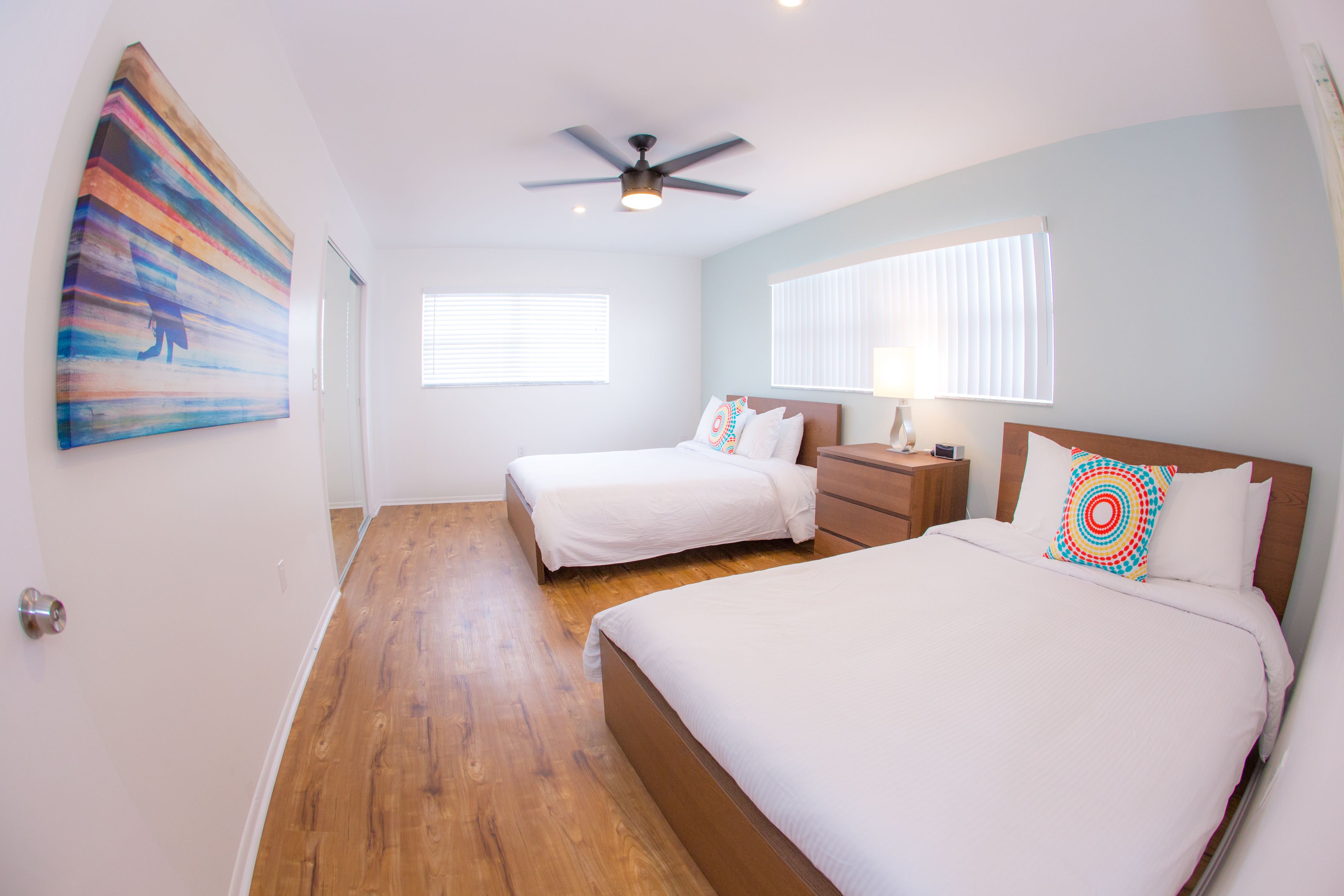 A hotel room with two beds, a ceiling fan, a painting on the wall, and a window with blinds.
