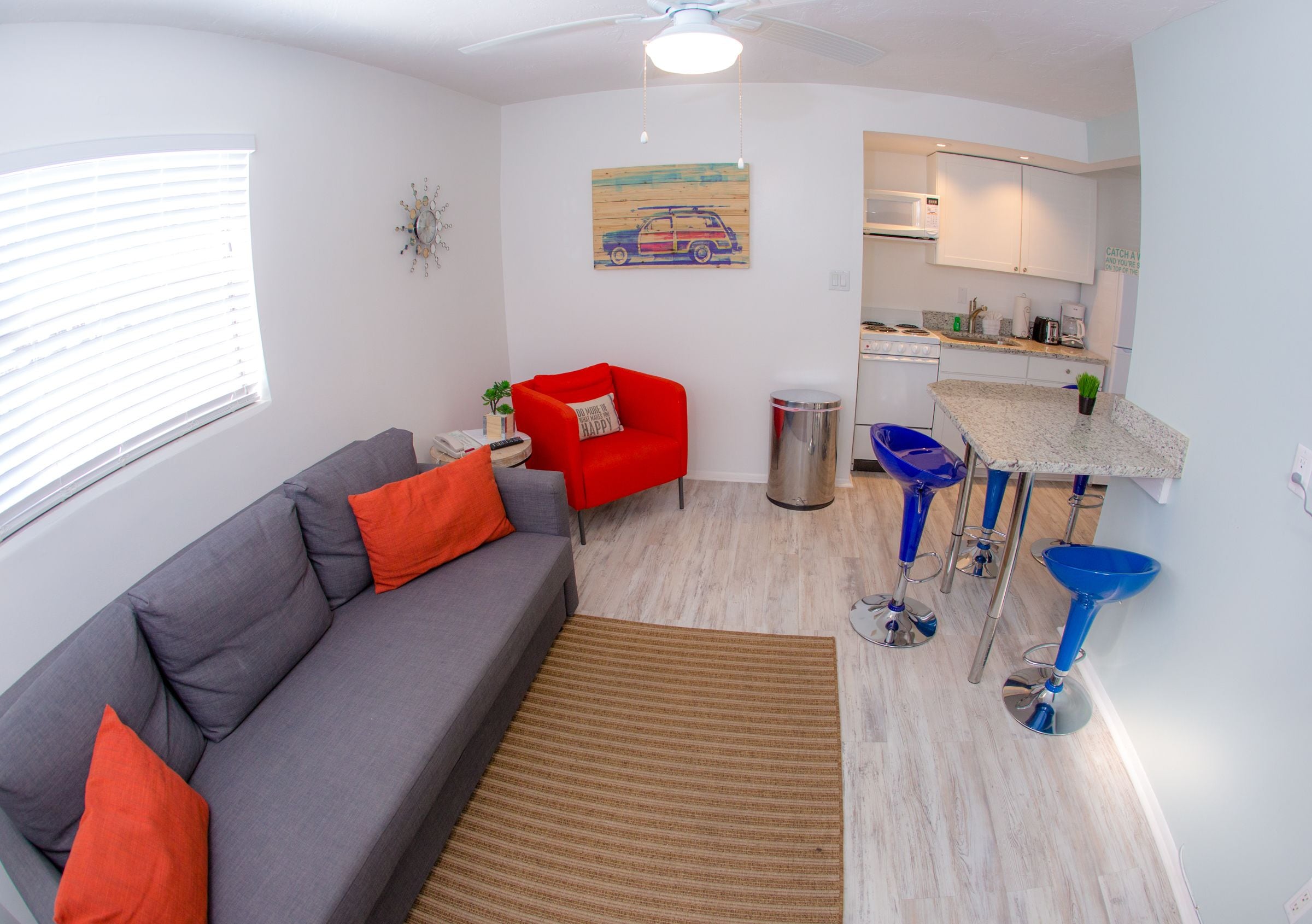 A compact living space with a grey sofa, red accent chair, kitchen area, and a dining table with blue bar stools.