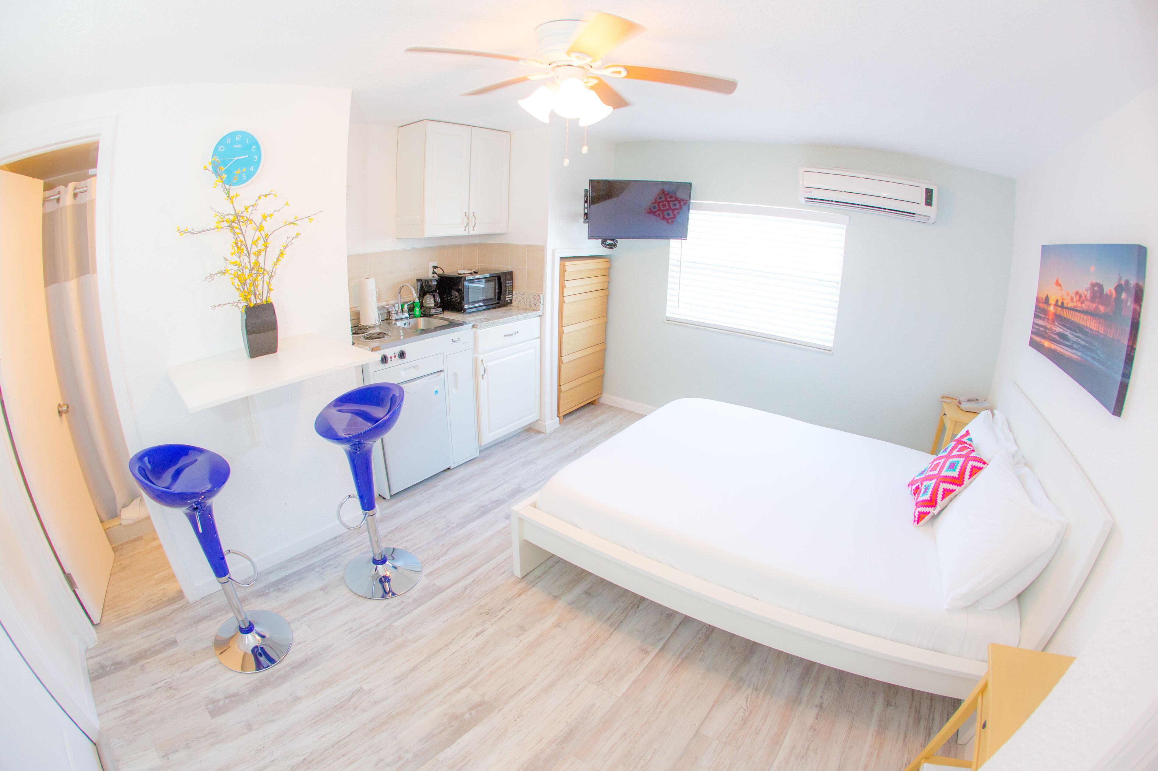 A compact bedroom with a bed, a ceiling fan, a TV, and a small kitchenette.