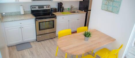 Cocina pequeña con armarios blancos, electrodomésticos de acero inoxidable, una mesa de madera con sillas amarillas y un letrero motivacional en la pared.
