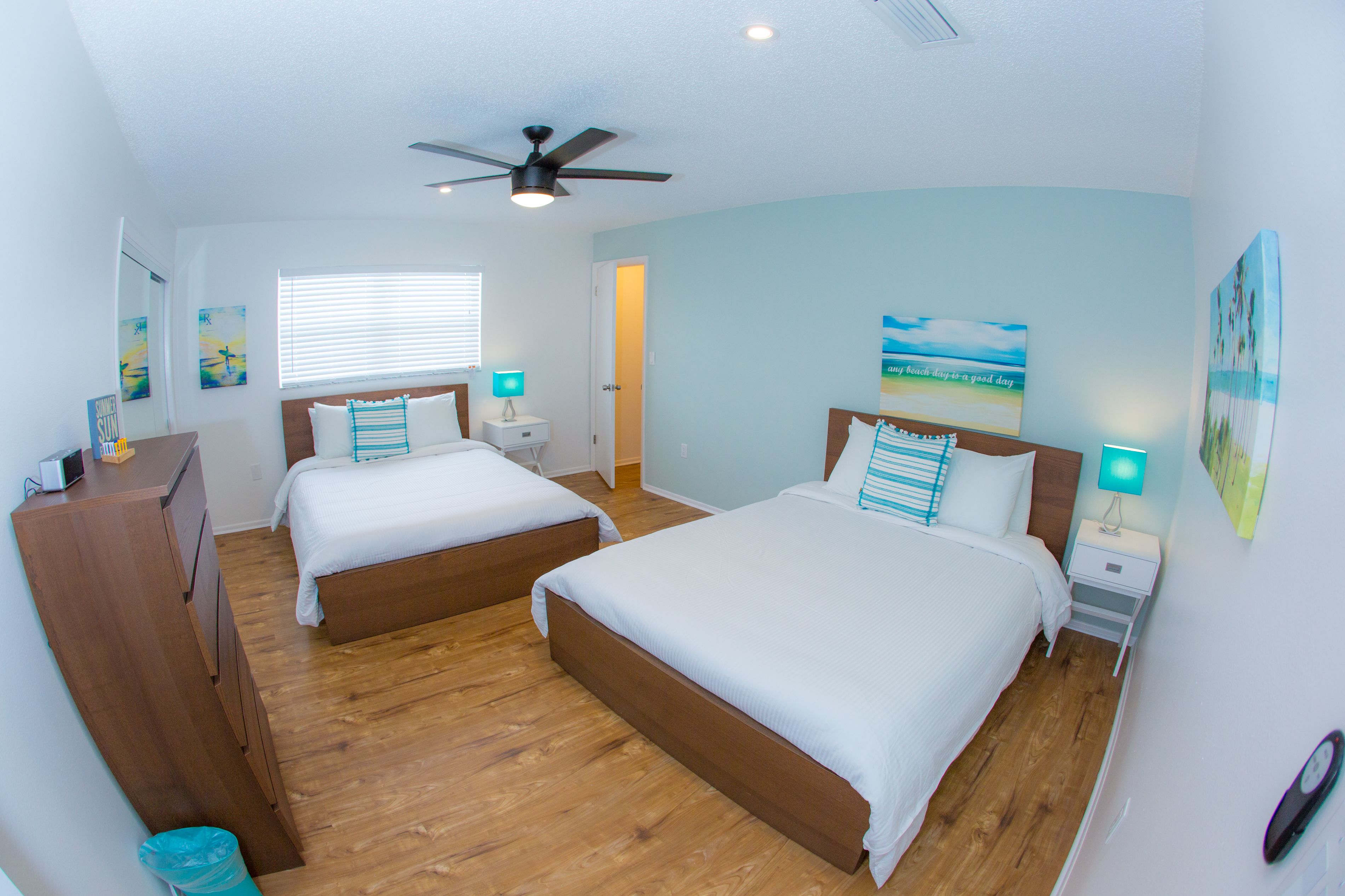 A hotel room with two beds, wooden flooring, a ceiling fan, a window with blinds, and artwork on the walls.