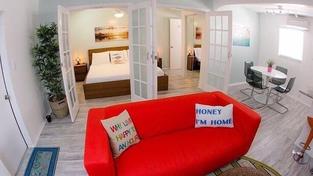 A modern hotel room with a red sofa, a bed with white bedding, a dining area with a table and chairs, and a large window. - St Pete Beach Suites (St. Pete Beach)