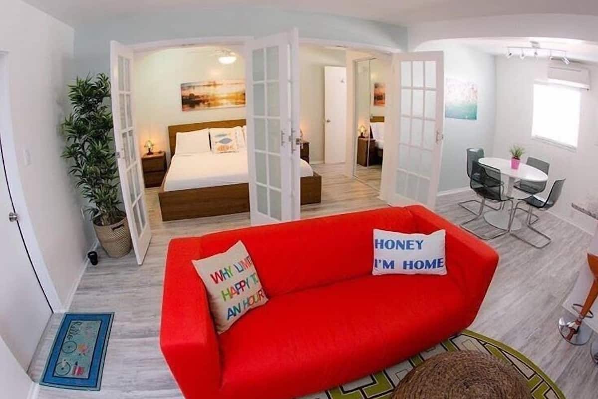 Une chambre d’hôtel moderne avec un canapé rouge, un lit avec une literie blanche, un coin repas comprenant une table et des chaises, et une grande fenêtre.