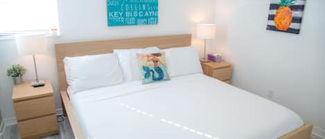 Una cama bien hecha con sábanas blancas, un cabecero de madera y dos mesitas de noche con lámparas. Se ve una obra de arte de colores y un cuadro de una piña.