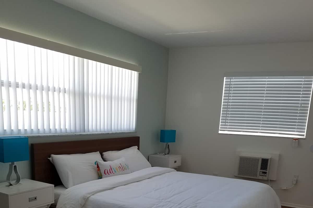 Une chambre bien rangée, avec un lit, des tables de chevet, une lampe et une fenêtre équipée de stores.