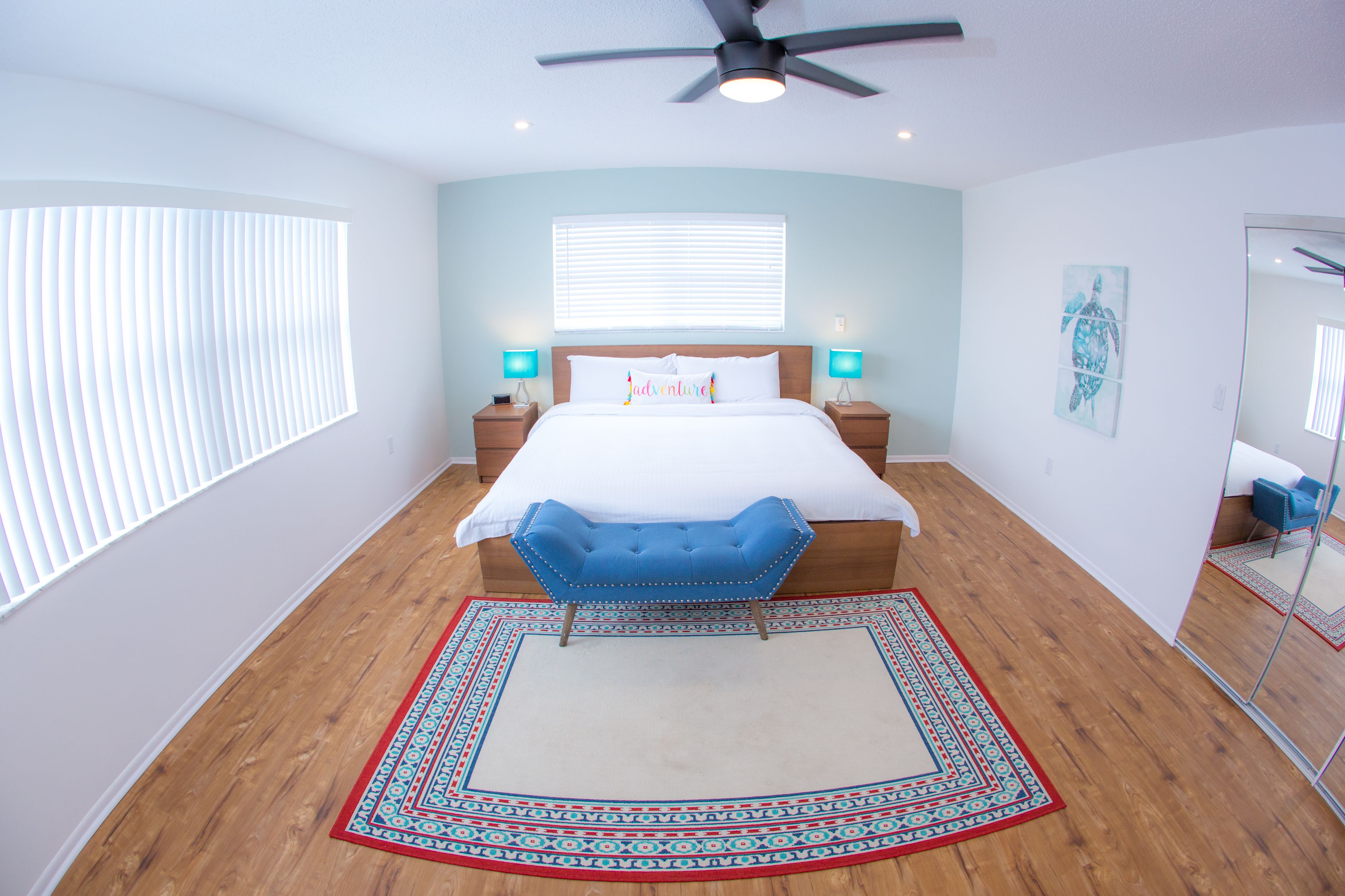 Une chambre à coucher avec un lit, une chaise bleue, un ventilateur de plafond, des lampes de chevet, une fenêtre avec des stores et un miroir.