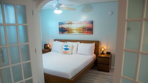 A bedroom with a bed, a nightstand, a ceiling fan, and a wall-mounted painting. - St Pete Beach Suites (St. Pete Beach)