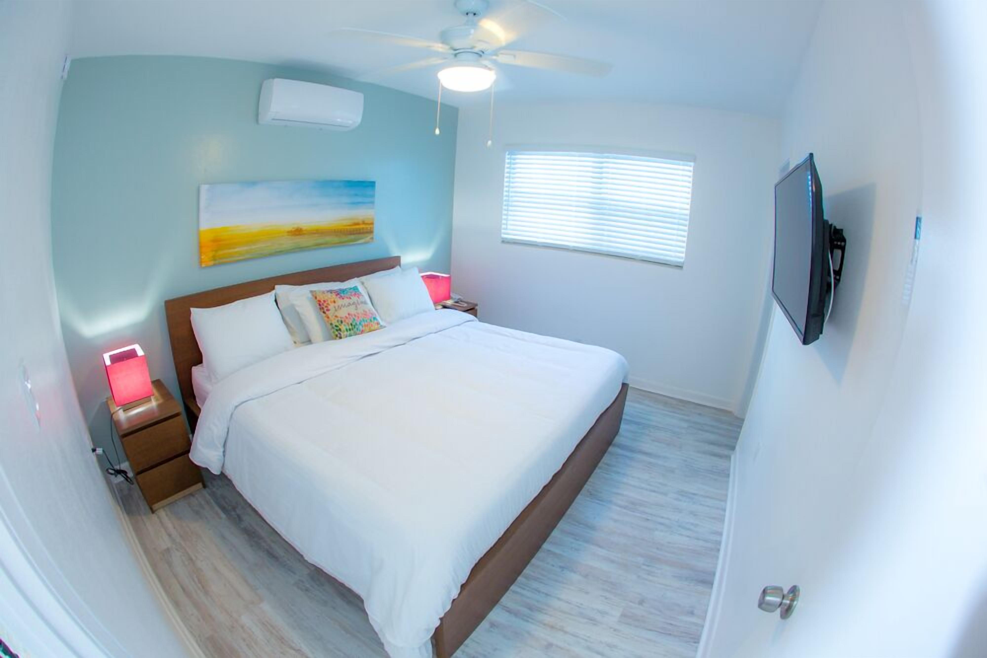 Une chambre à coucher comprenant un lit, des tables de chevet, un ventilateur de plafond, une télévision et une fenêtre avec des stores.