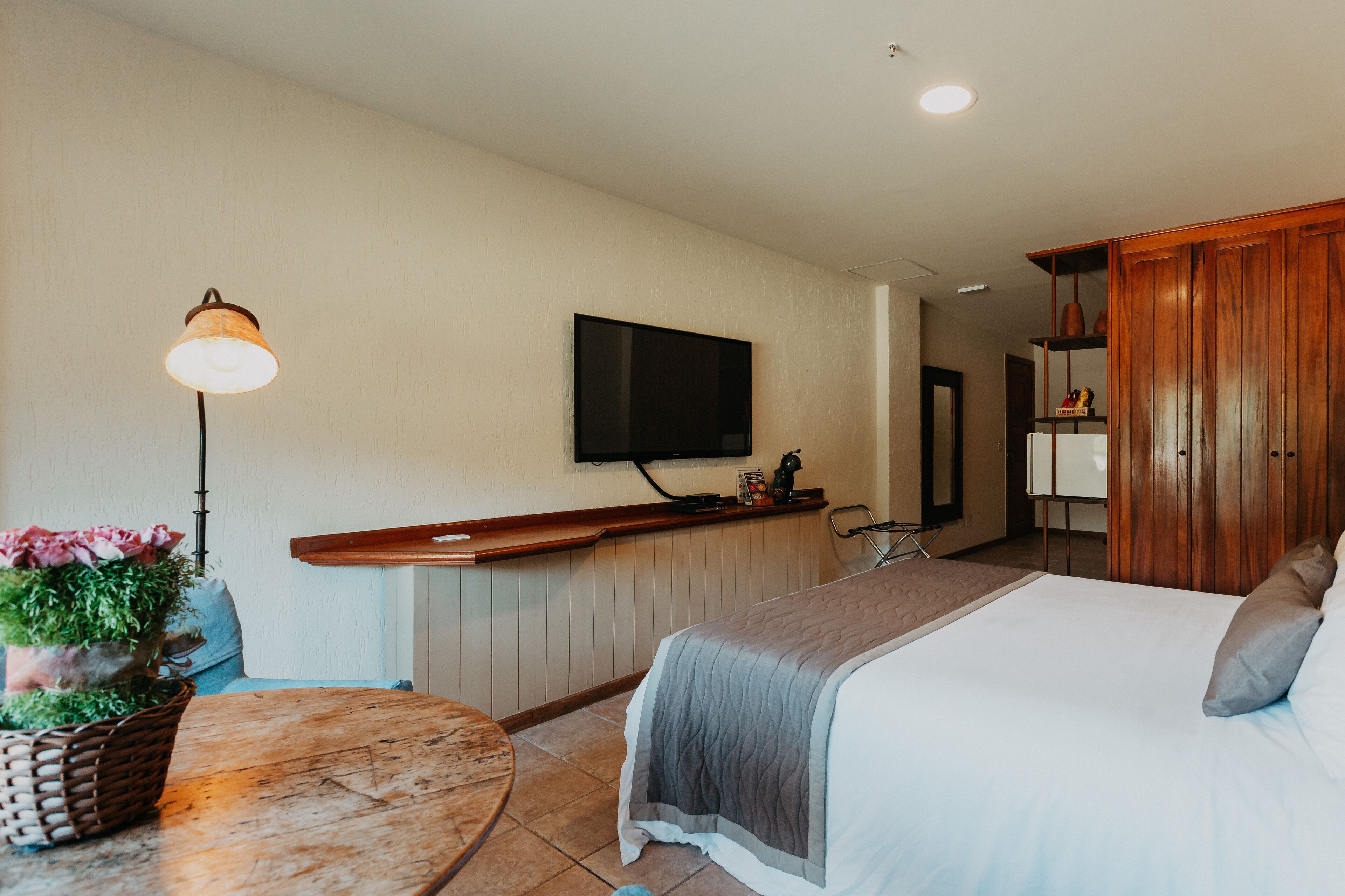 A hotel room with a bed, a television mounted on the wall, a wooden cabinet, and a small table with a lamp and a plant.