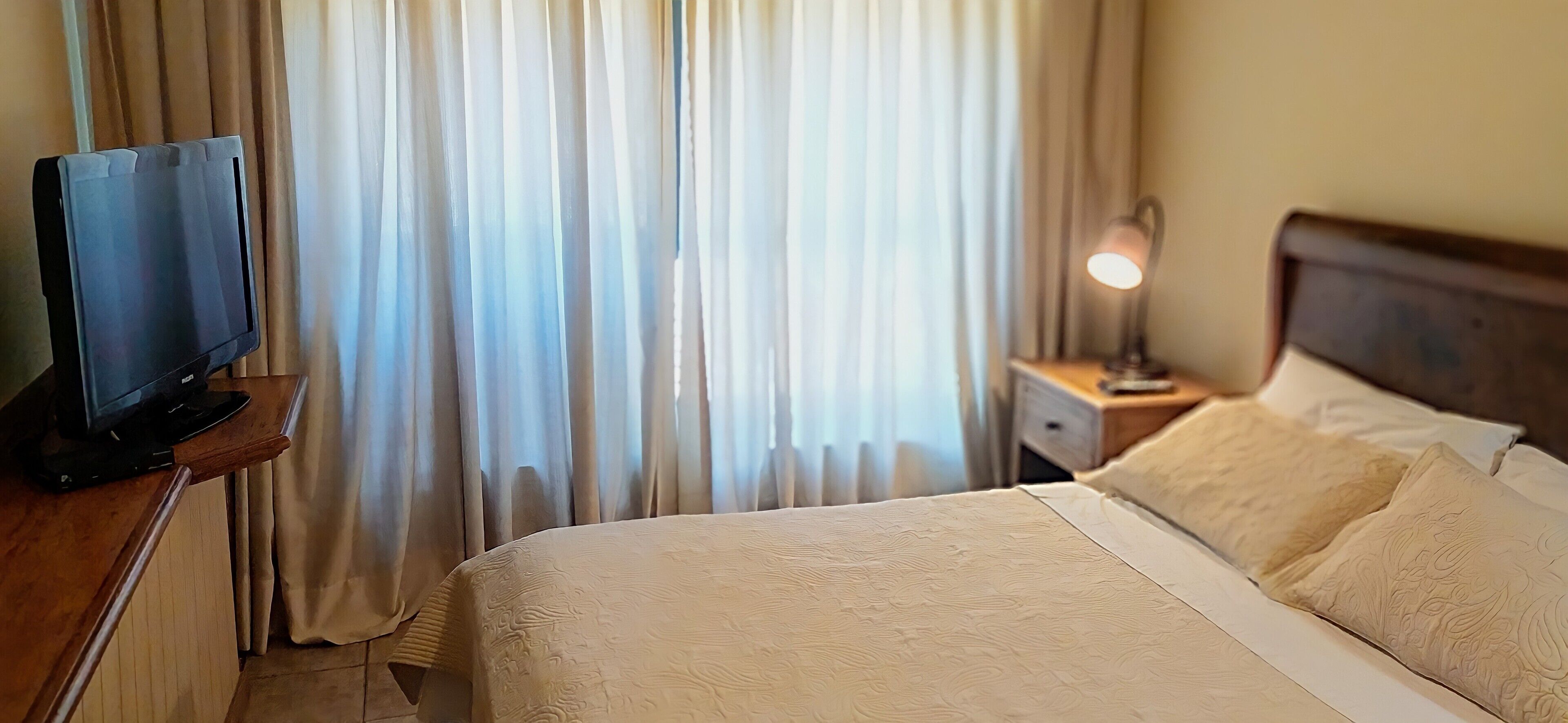 Habitación de hotel con cama, televisor sobre un soporte de madera, mesita de noche con lámpara y ventana con cortinas.