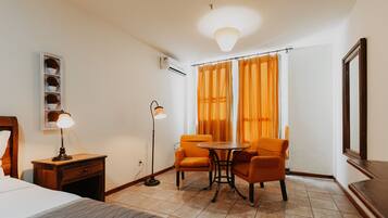 Une chambre d’hôtel avec un lit, un bureau doté d’une lampe, deux chaises disposées autour d’une petite table, un appareil de climatisation et une fenêtre avec des rideaux orange.