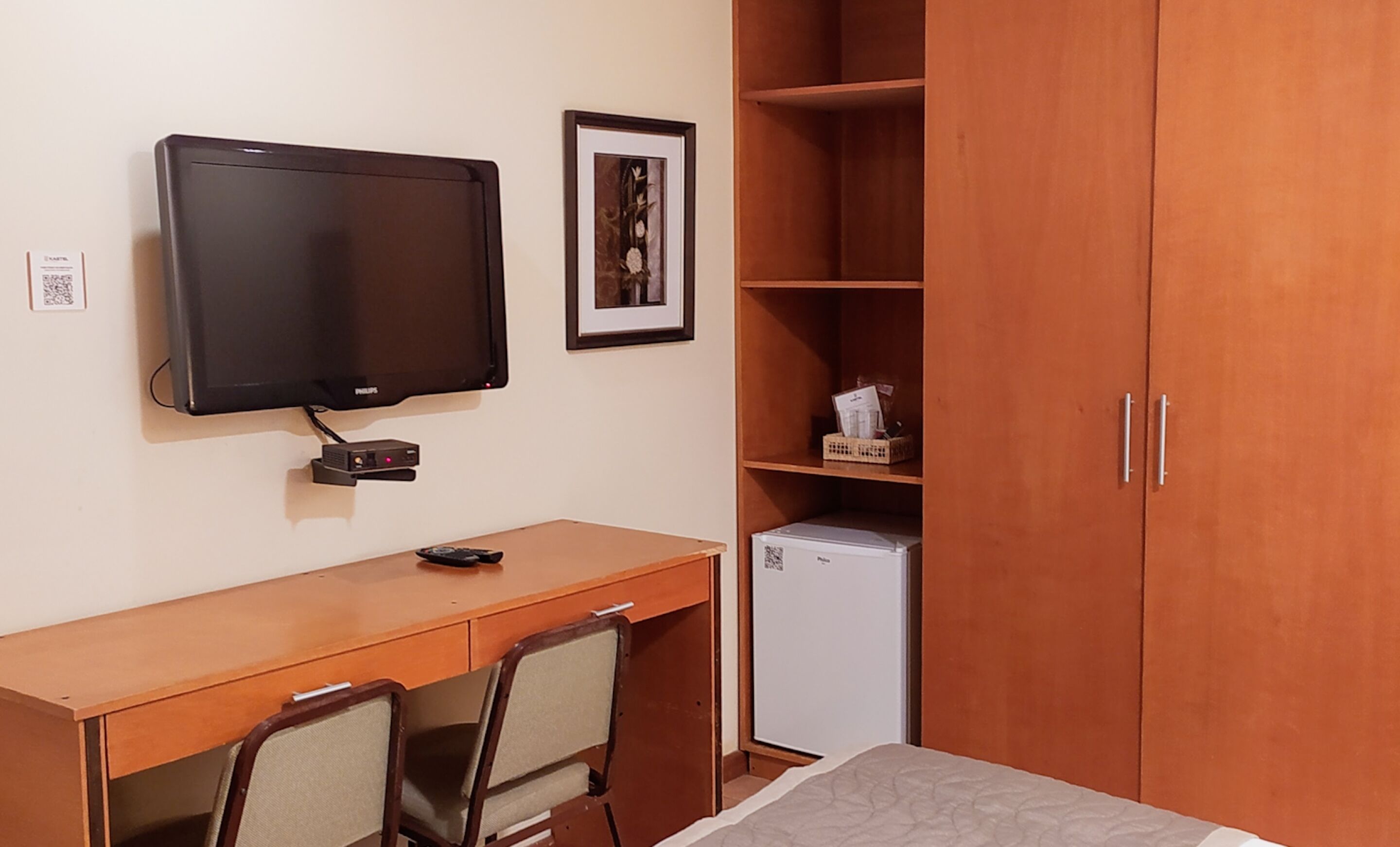 Une chambre avec un bureau en bois, une télévision, un petit réfrigérateur et une armoire.