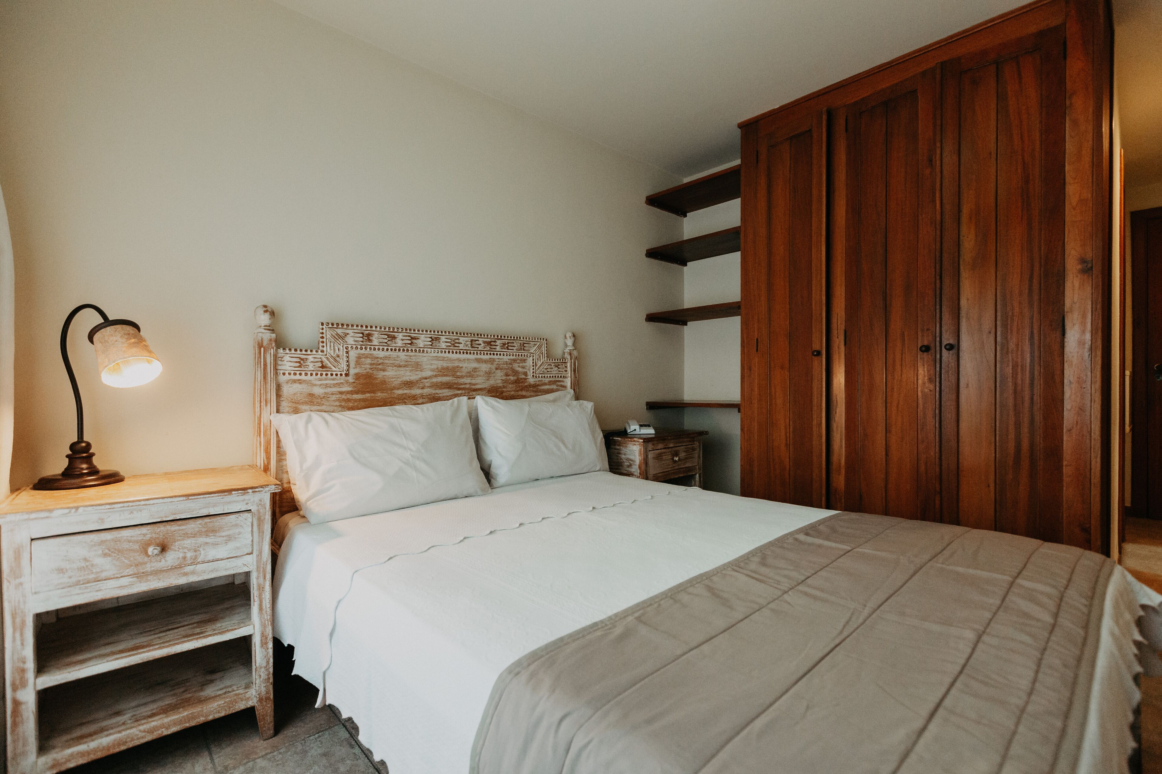 Un dormitorio con cabecera de madera, mesita de noche con lámpara, armario y cama con ropa blanca.