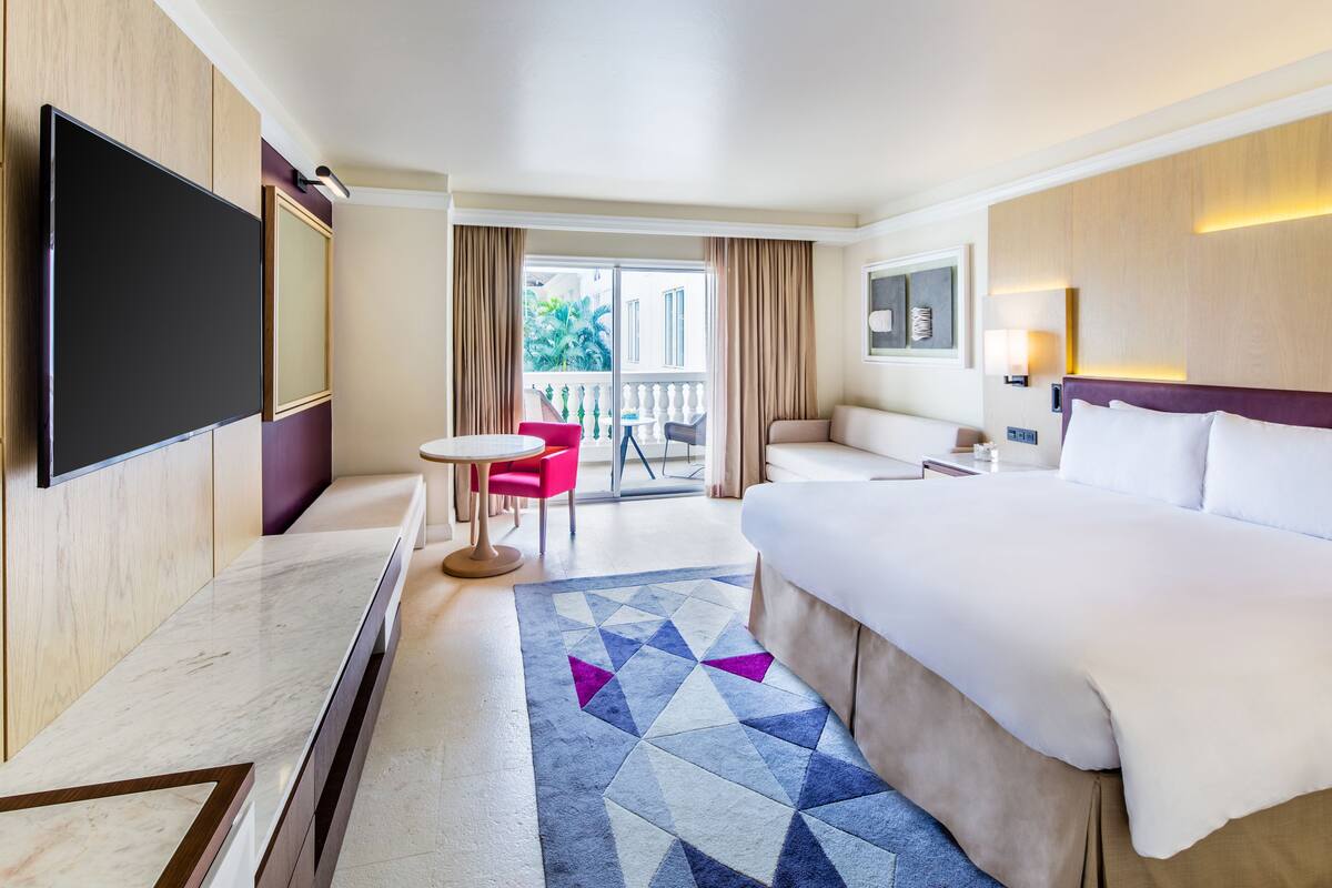 A hotel room with a large bed, a flat-screen TV mounted on the wall, a small round table, a red chair, and a balcony with a view of greenery.
