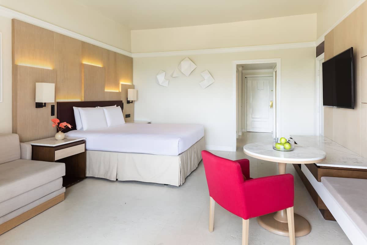 A hotel room with a bed, a desk with a chair, a small table with fruit, and a TV on the wall.