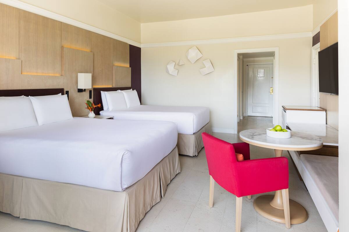 A hotel room with two beds, a small round table with a bowl of fruit, a red chair, and a TV on the wall.