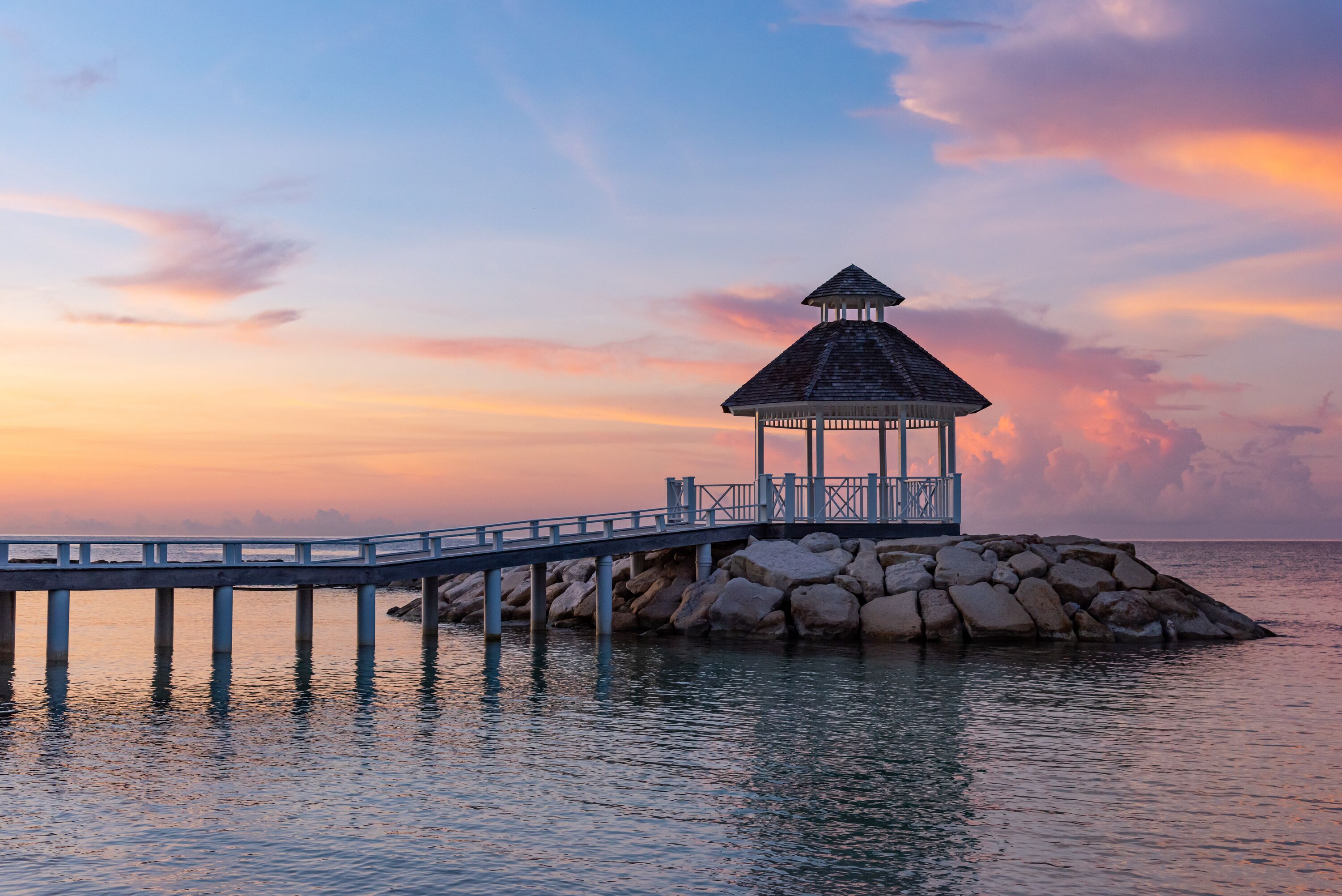 Ein Pavillon auf einem Steg, der sich über ruhiges Wasser erstreckt, mit einem Sonnenuntergangshimmel im Hintergrund.