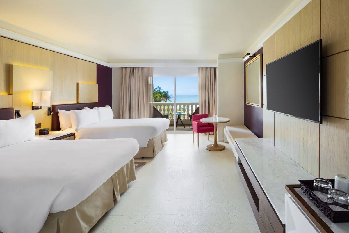 A hotel room with two beds, a TV, a small table, and a balcony with a view of the ocean.