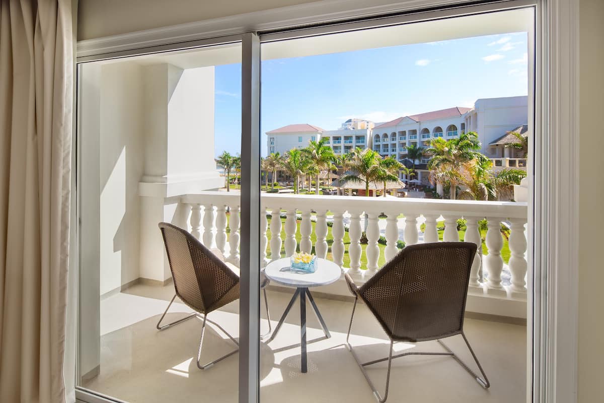A balcony with a glass door, two wicker chairs, a small round table, and a view of a hotel building with palm trees.