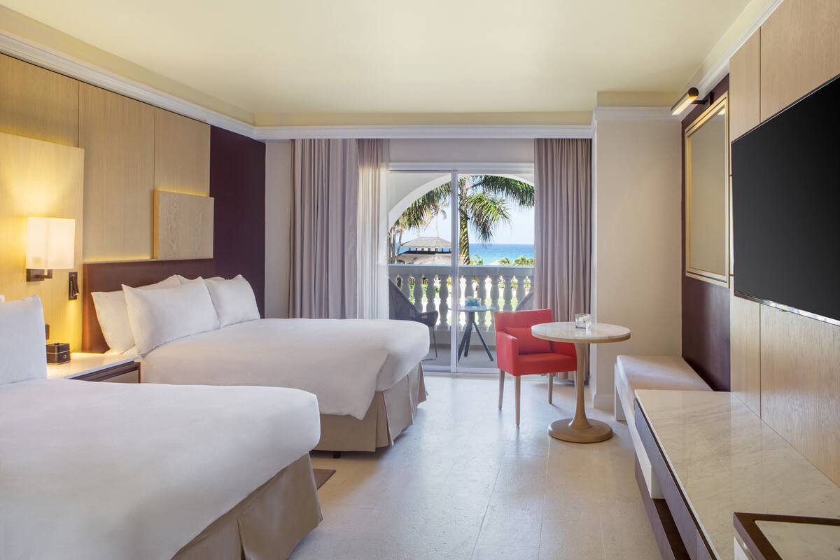 A hotel room with two beds, a small table, a TV, and a balcony with a view of the ocean.