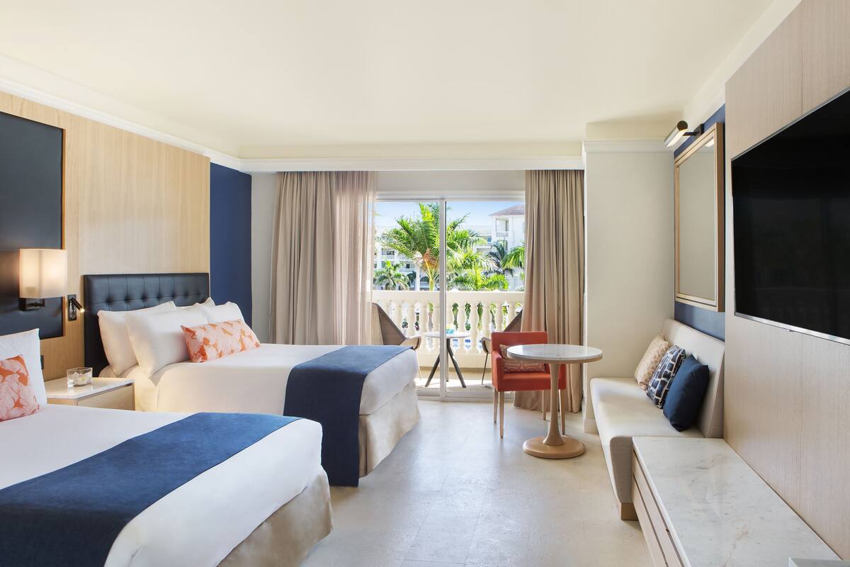 A hotel room with two beds, a sofa, a small table, and a balcony with a view of palm trees.