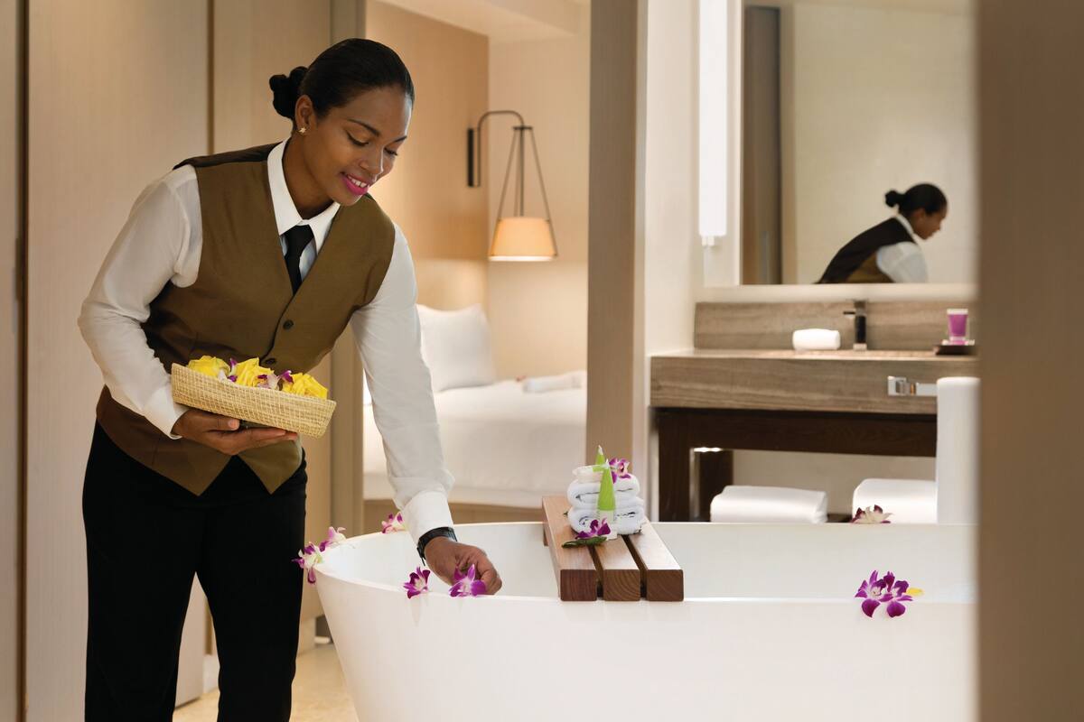 A hotel room with a white bathtub, a wooden side table with towels and toiletries, and a basket of towels and flowers on the floor.