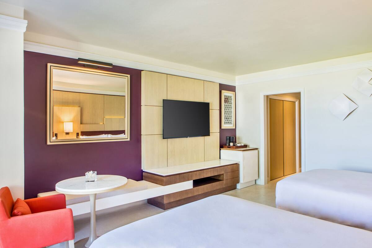 A modern hotel room with a large bed, a TV mounted on the wall, a small table, and a red chair.