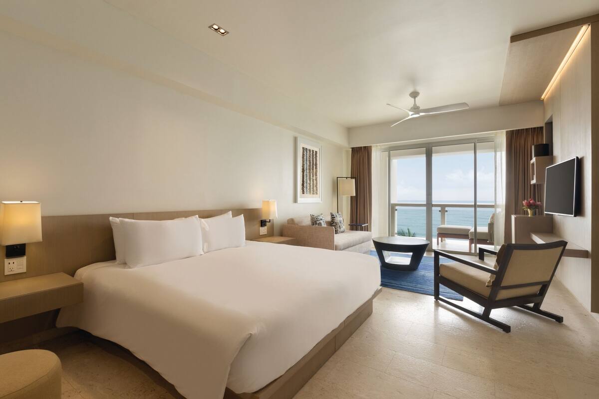 A hotel room with a large bed, a desk, a chair, a TV, and a balcony with a view.