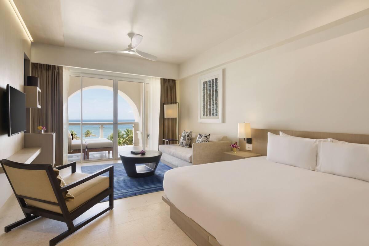 A hotel room with a large bed, a desk, a chair, a sofa, a balcony with a view of the ocean, and a ceiling fan.