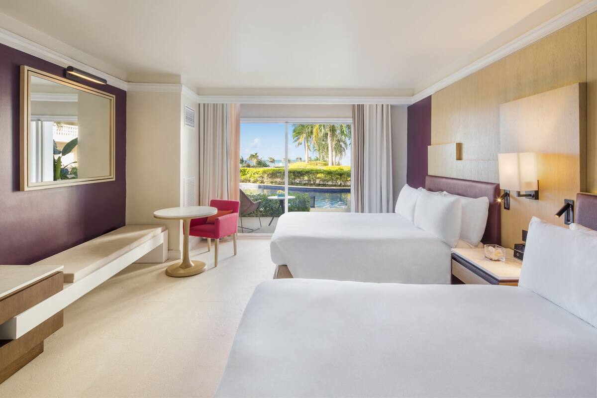 A hotel room with two beds, a small table, a chair, a mirror, and a view of a pool and palm trees.