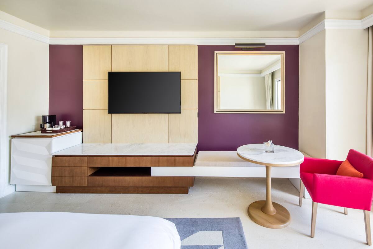 A modern hotel room with a bed, a TV mounted on the wall, a small round table, a red chair, and a large mirror.