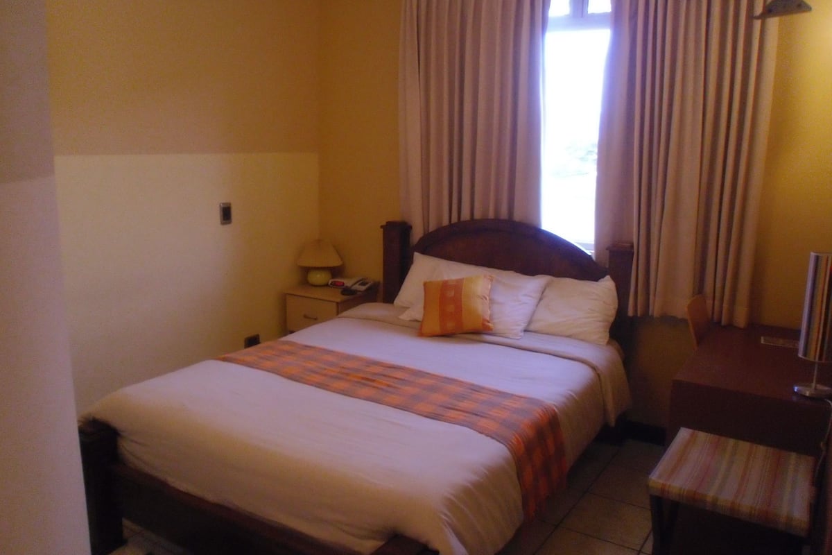 A hotel room with a bed, a nightstand, a lamp, a chair, and a window with curtains.